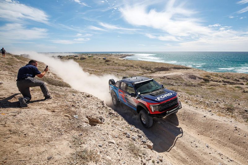 Person with cell phone on one knee videoing the take off of a Ford branded performance truck on desert ground and the ocean in the background